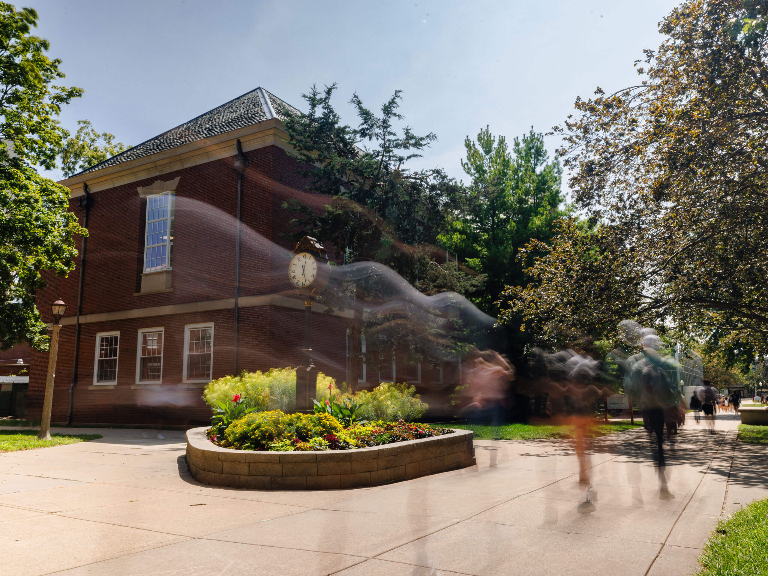 A red-brick building with a vintage clock and blurred figures walking by a flower bed.