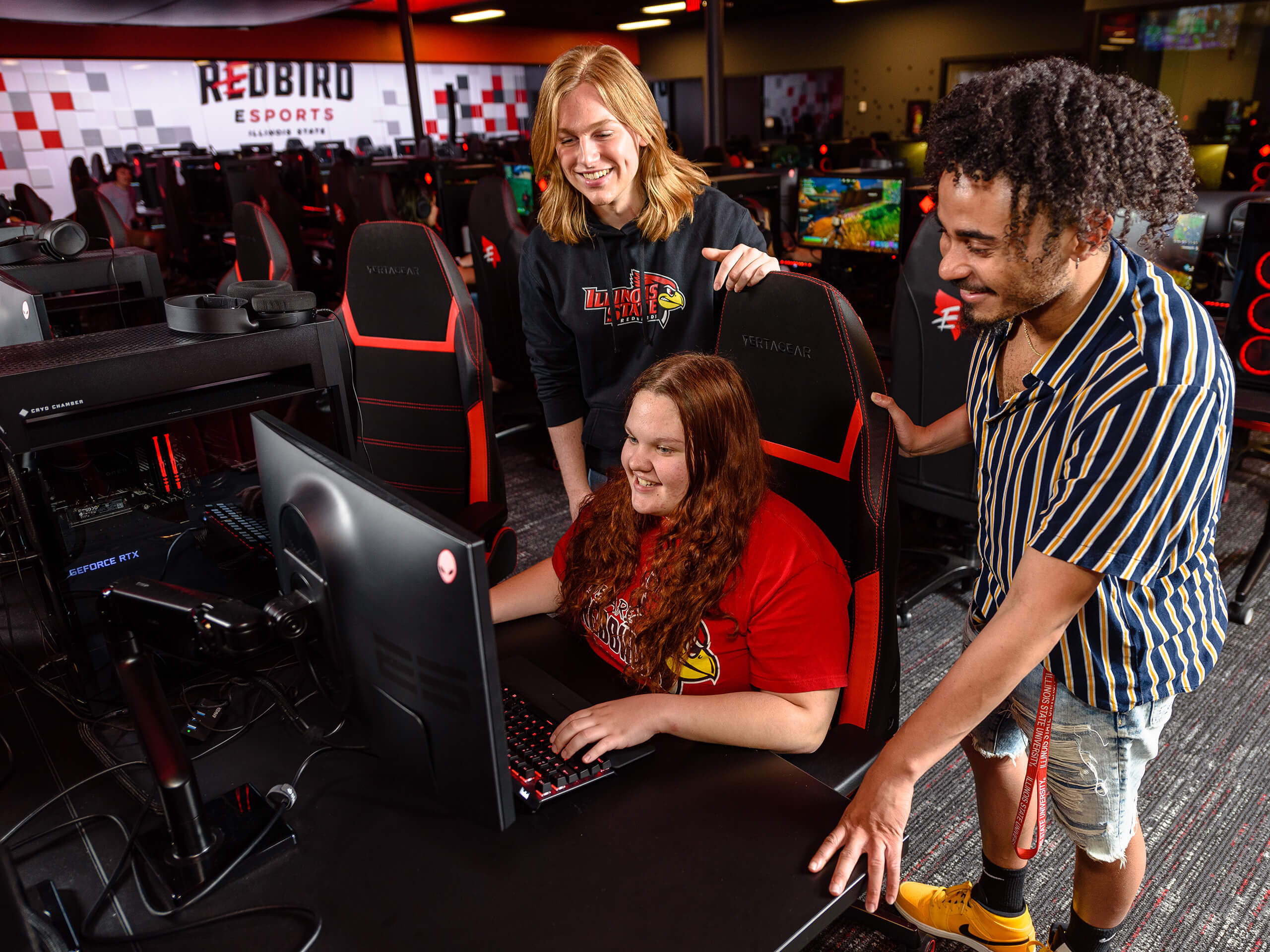 Three people in an esports center, one seated at a computer, two standing beside them.