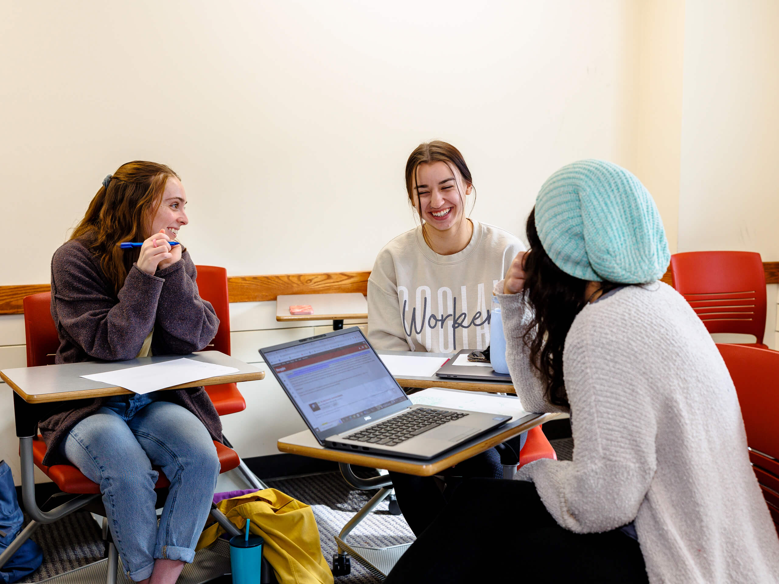Three students in a classroom sitting in red chairs, engaged in a lively discussion.