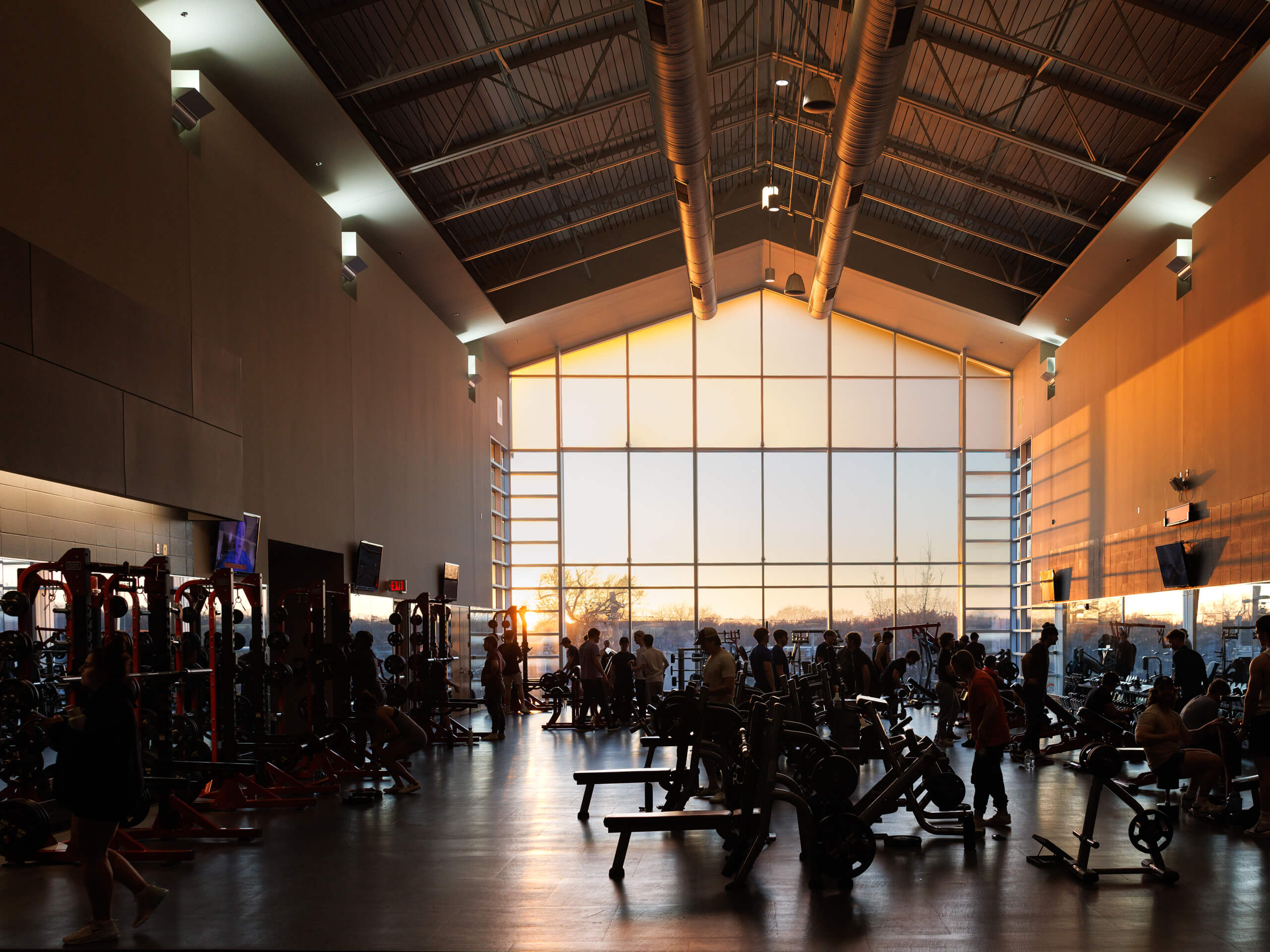 Gym interior at sunset with people using exercise equipment, lit by golden sunlight from large windows.