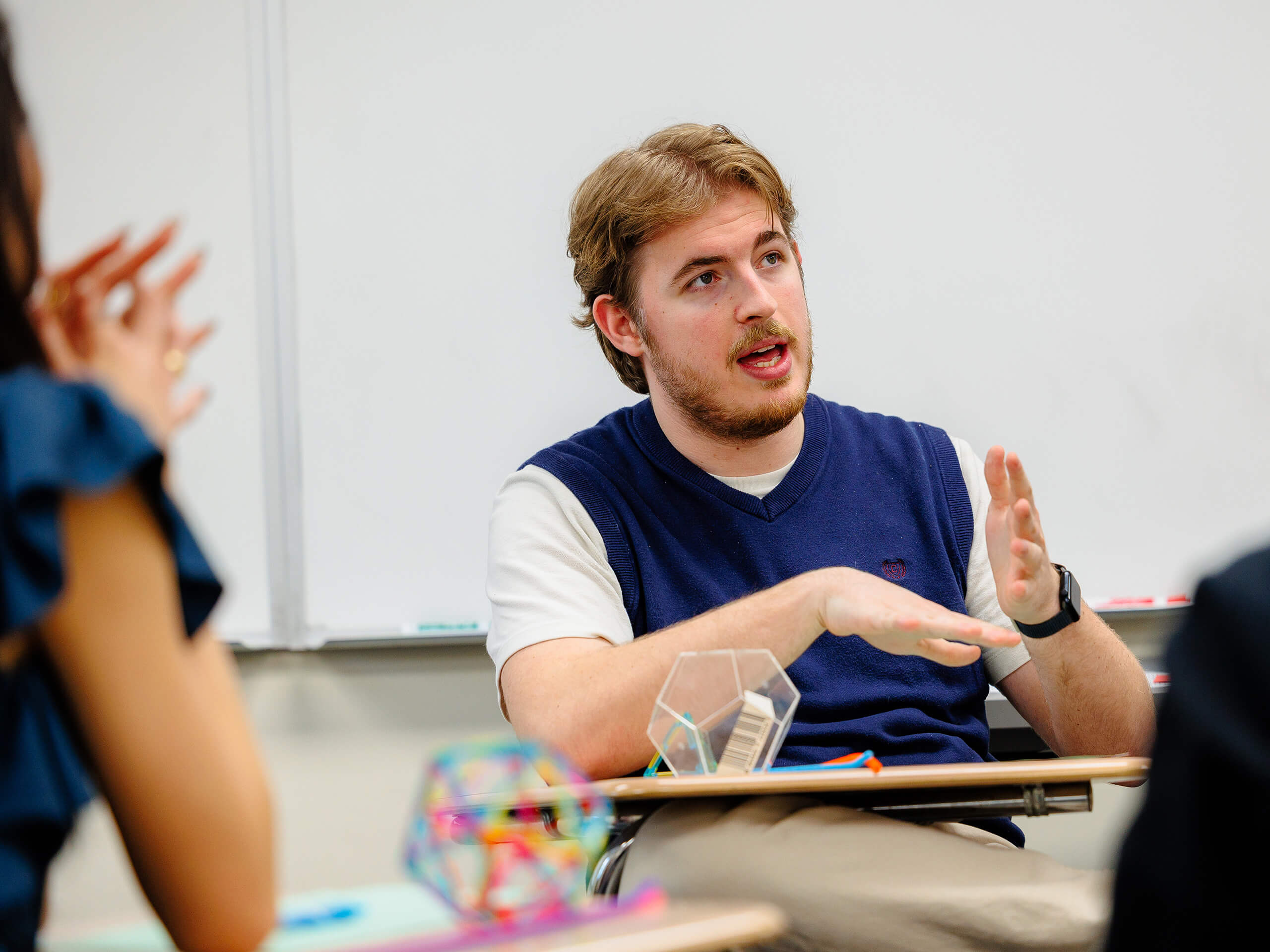 A person gestures while speaking in a classroom, with geometric models on the desk in front.