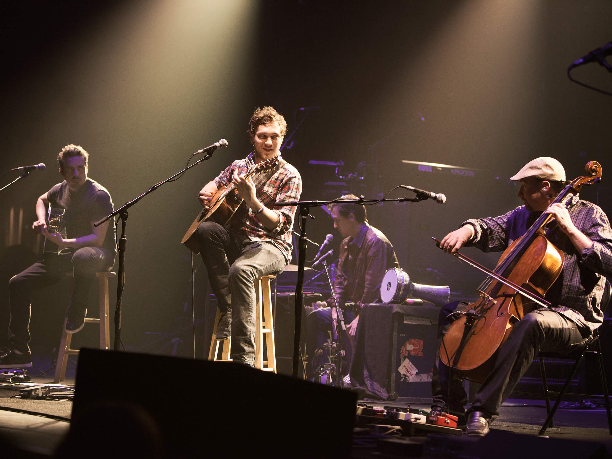 Four musicians perform on stage, including two guitarists, a cellist, and another musician playing a keyboard.