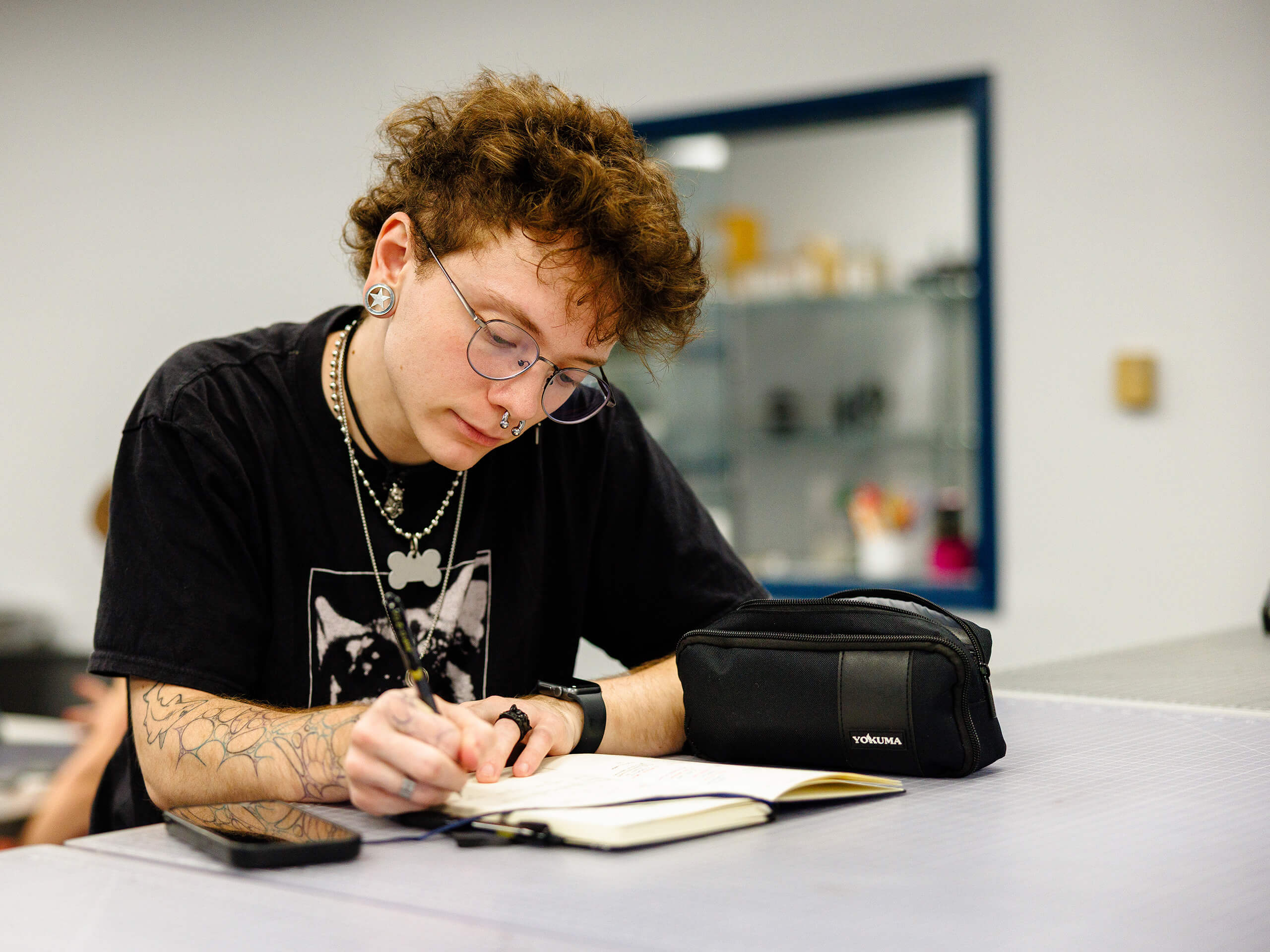 Person with curly hair writing in a notebook at a table, with a pencil case beside them.