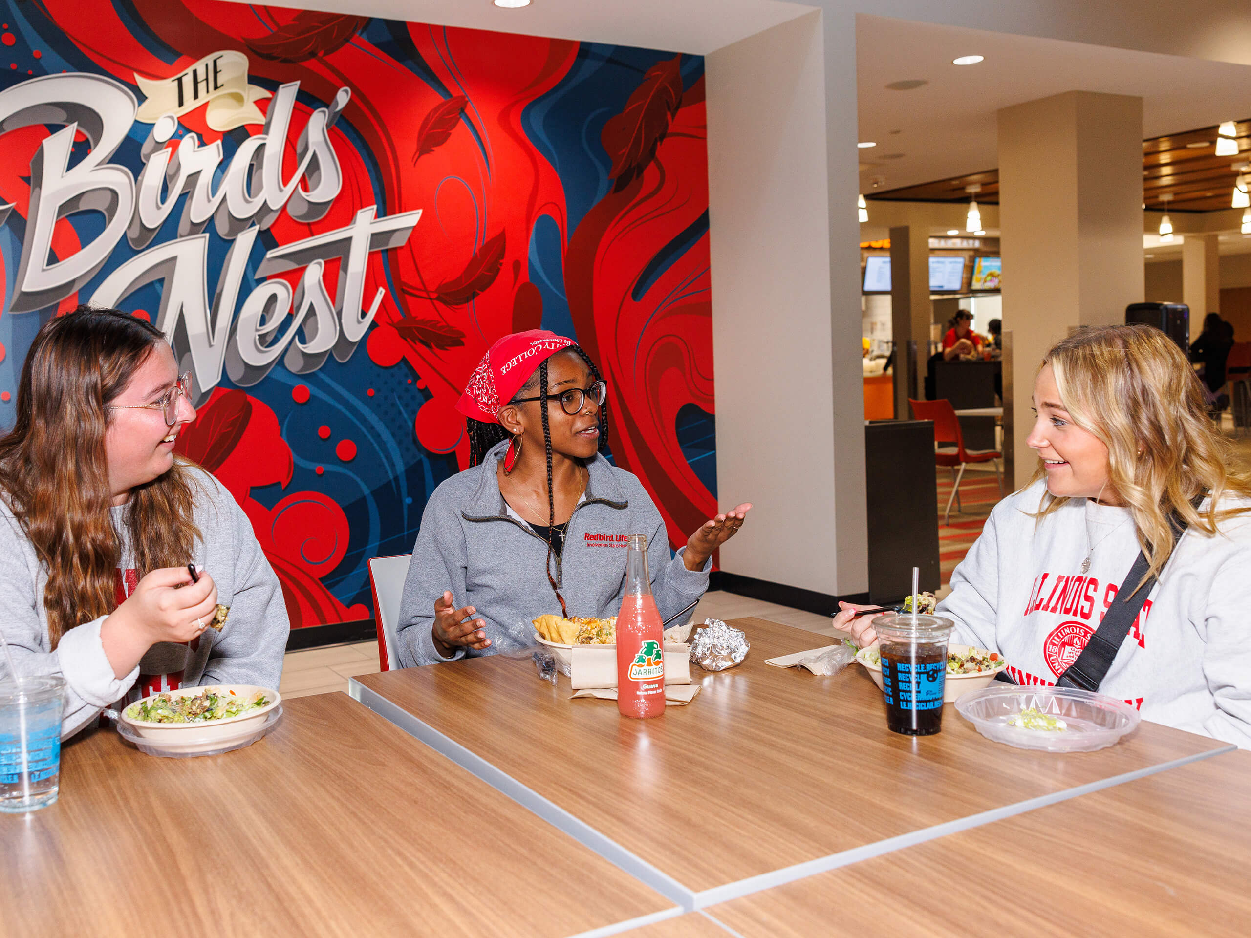 Three people sitting at a table in front of "The Bird's Nest" mural, engaged in conversation.