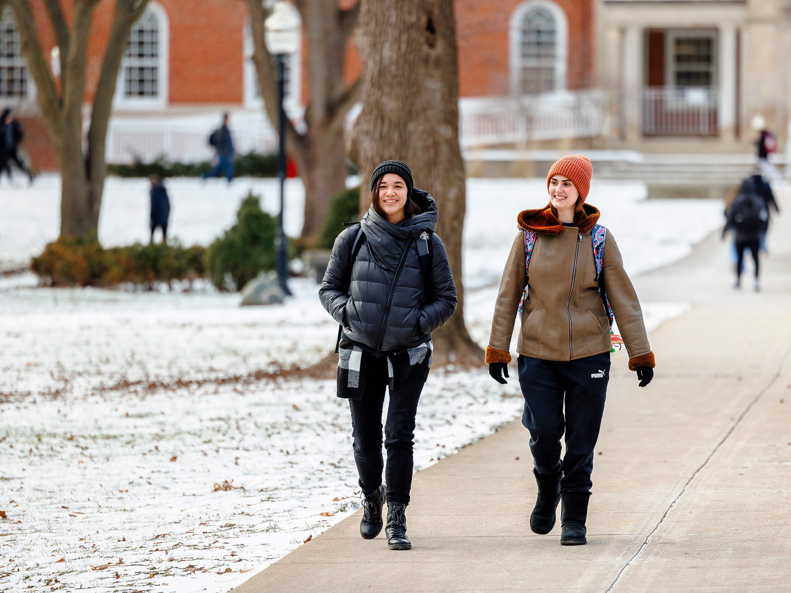 Two people walking on a snowy college campus path, wearing winter clothes.
