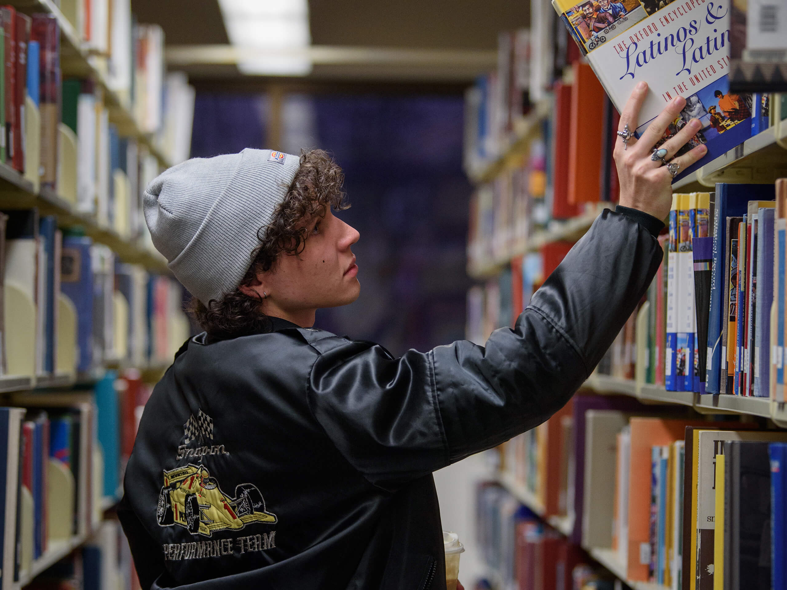 Person in a library reaching for a book on a shelf, wearing a gray beanie and black jacket.