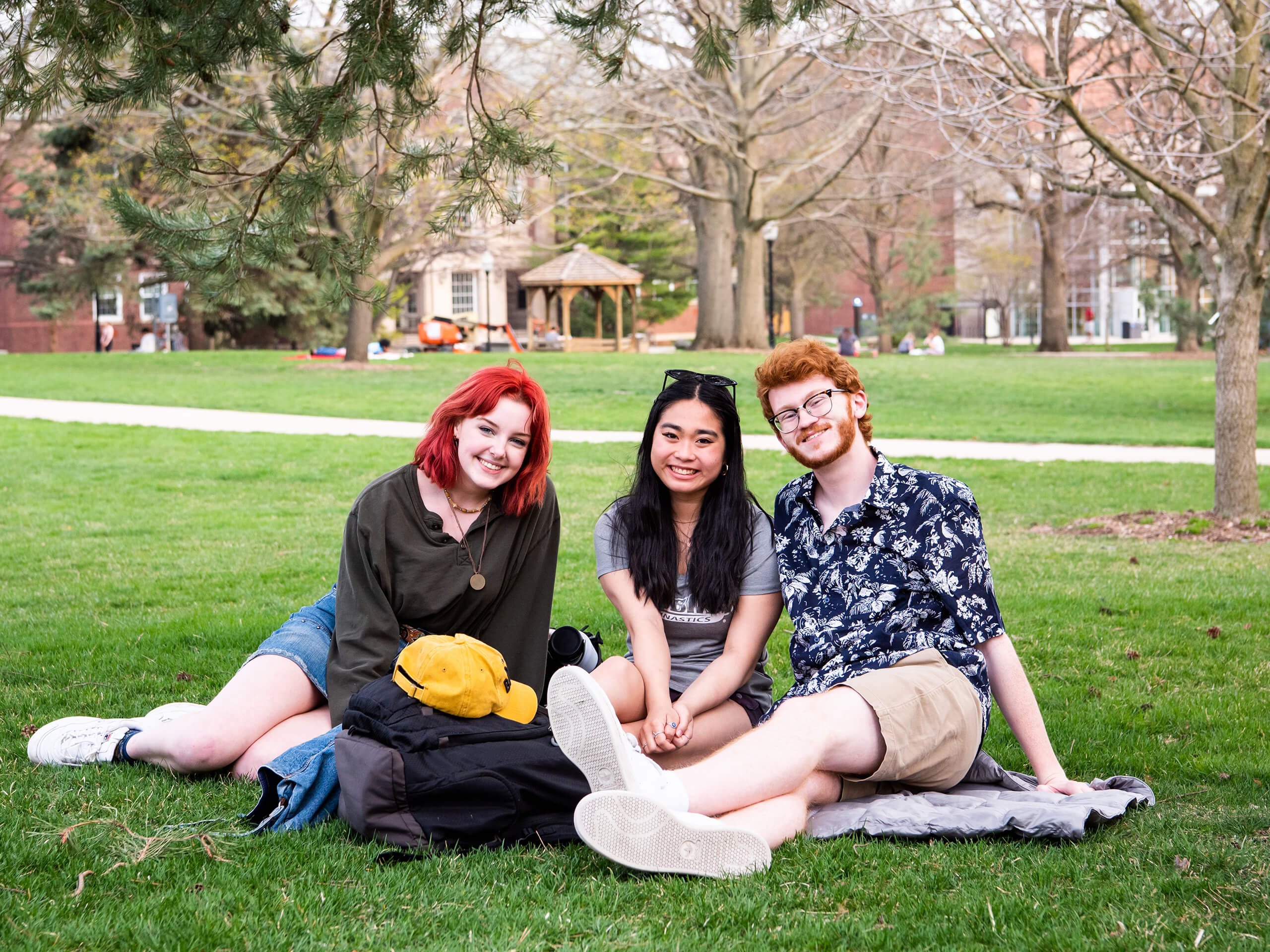 Three students sit on the quad together, with buildings and trees in the background.