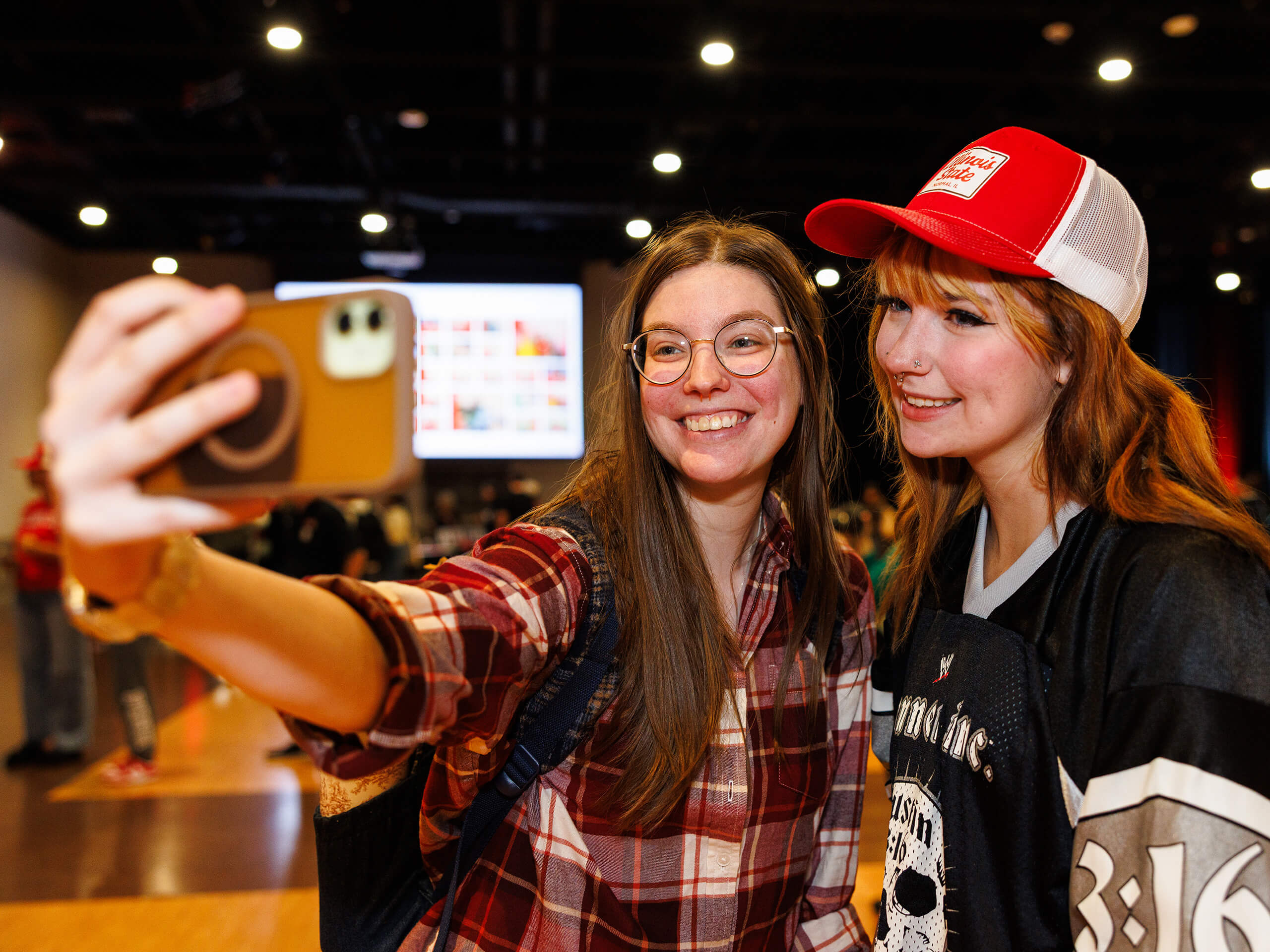 Two people taking a selfie indoors, one wearing a red plaid shirt and glasses, the other in a red hat and black shirt.
